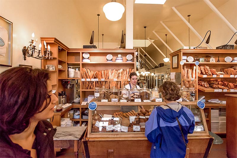 People standing at the counter of a bread shop, where you can see bread in baskets piled high