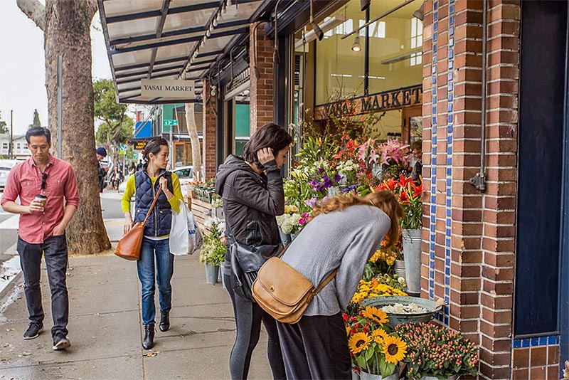 Two people smell flowers outside of a shop while two other people walk past the shop, looking in the window