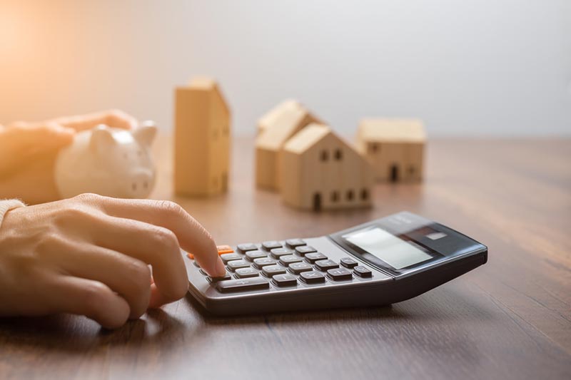 A hand pressing a calculator beside a piggy bank and small, wooden houses