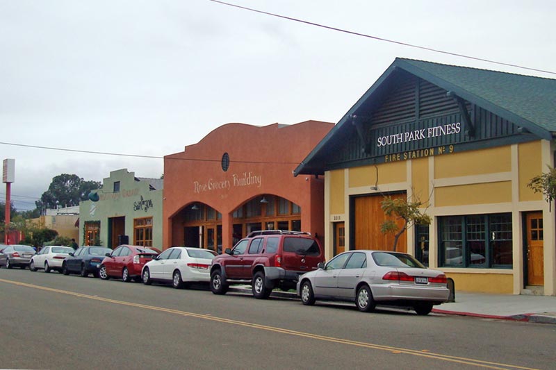 Row of businesses along neighborhood street with cars parked outside.