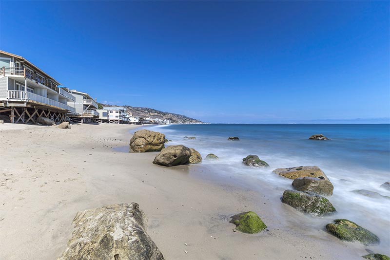 Water washes over rocks along the beach in Malibu with large homes in the background