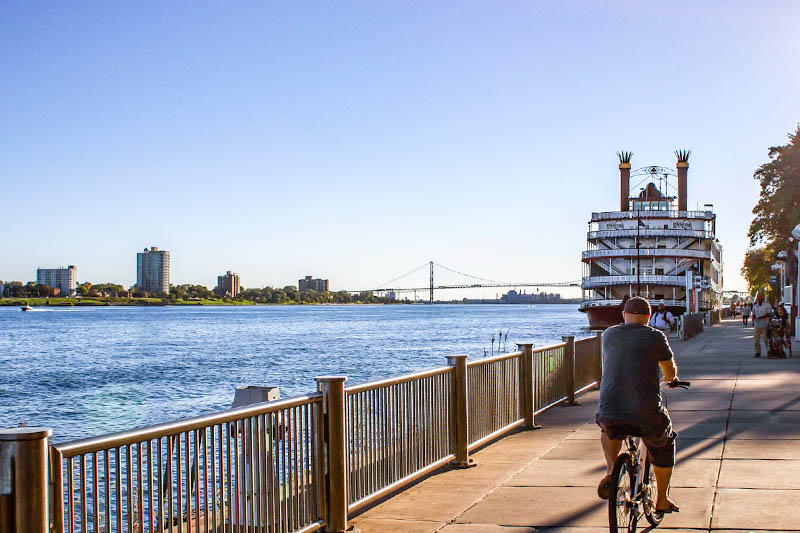 A man bikes down the riverwalk in Detroit with a boat in front of him on the river