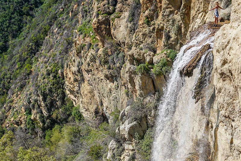 A person stands and poses at the top of Escondido waterfall as the falls rush down a canyon