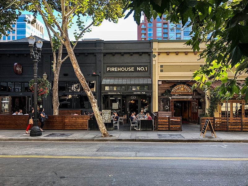 People eating and drinking on outdoor patio of shaded restaurant and people walking by on sidewalk.