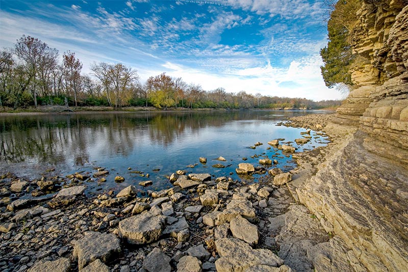 A rocky shoreline next to a river, and in the distance there's a line of trees at the Kankakee River outside Chicago