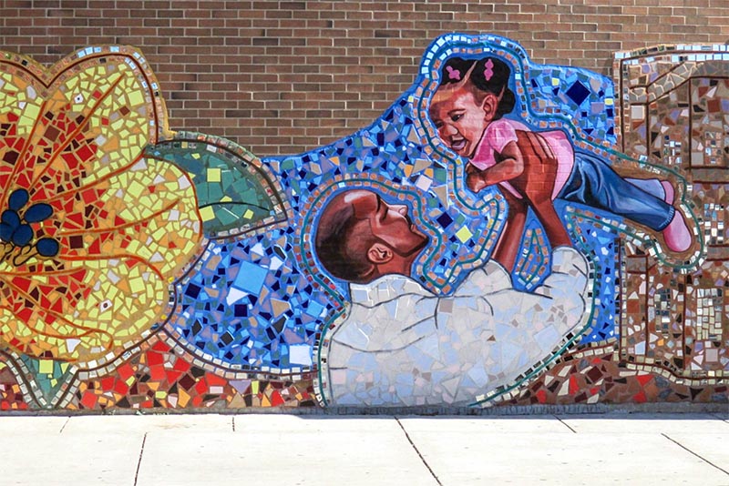 A mosaic mural depicts a man holding a child in the air surrounded by flowers