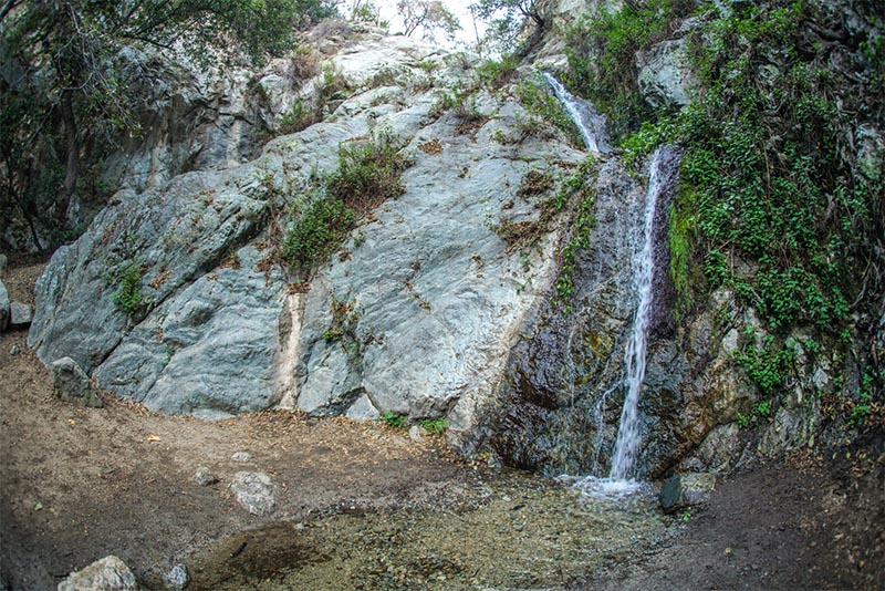 A thin waterfall flows over a number of large boulders before splashing onto the forest floor basin