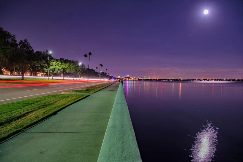 Night time view of pathway by the ocean and city skyline in the distance.