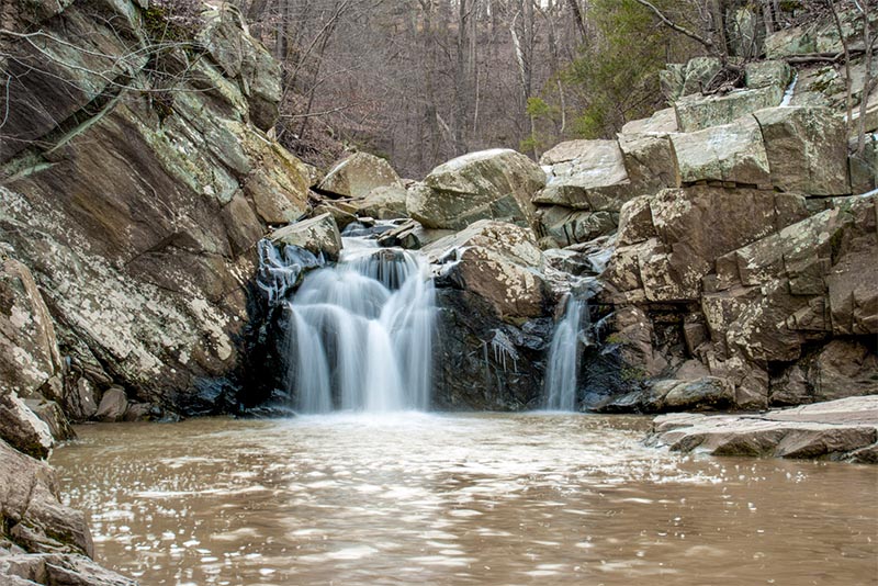 A short waterfall flows over large rocks surrounded by winter foliage