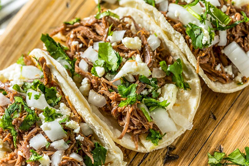 Three tacos on a wood board with meat, onion, and cilantro on them