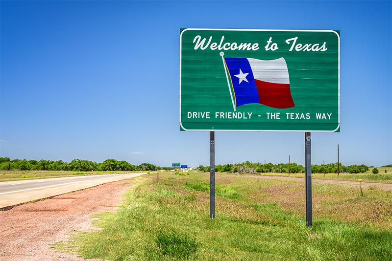 A green road sign along a two-lane highway that says Welcome to Texas with a Texas flag on it